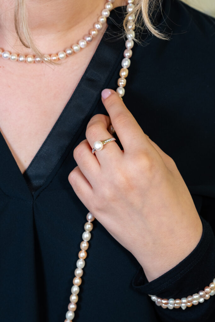 Nahaufnahme einer Hand, die eine Perlenkette festhält; am Finger trägt die Person einen Perlenring, kombiniert mit einem dunklen Oberteil.